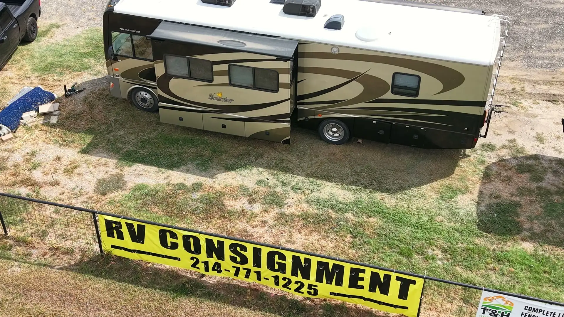 Yellow RV CONSIGNMENT sign with the phone number 214-771-1225 on the fence at the BK Consignments RV lot on I-30 in Greenville, Texas